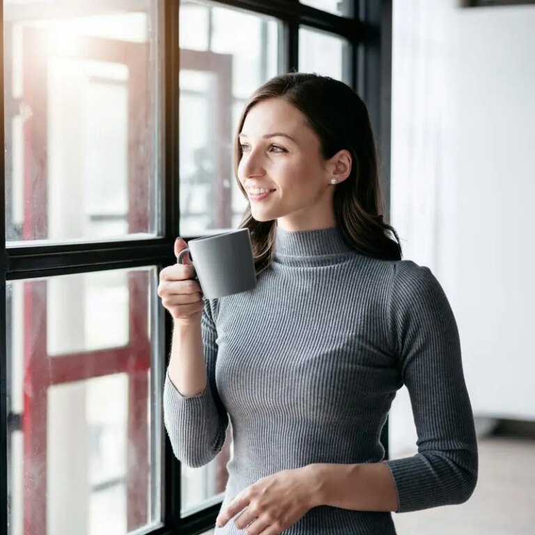 Businesswoman drinking hot chocolate and standing near window to looking outside for take a break